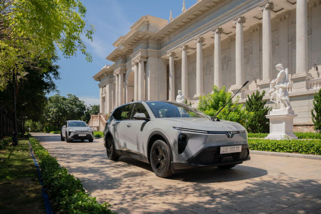 A grey VinFast VF MPV 7 leading a convoy on a paved driveway past classical European-style architecture.